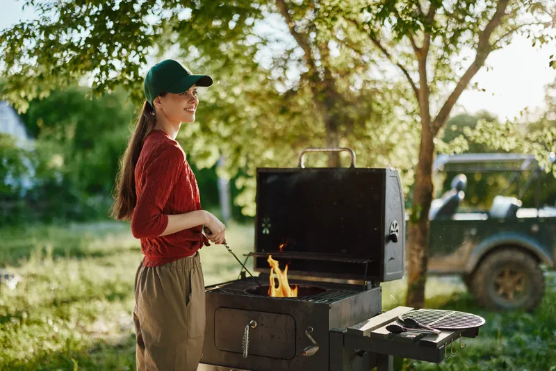 A young woman grilling outdoors in a nature setting, wearing a red shirt, green cap, and a relaxed expression, showcasing summer vibes and a joyful cooking moment