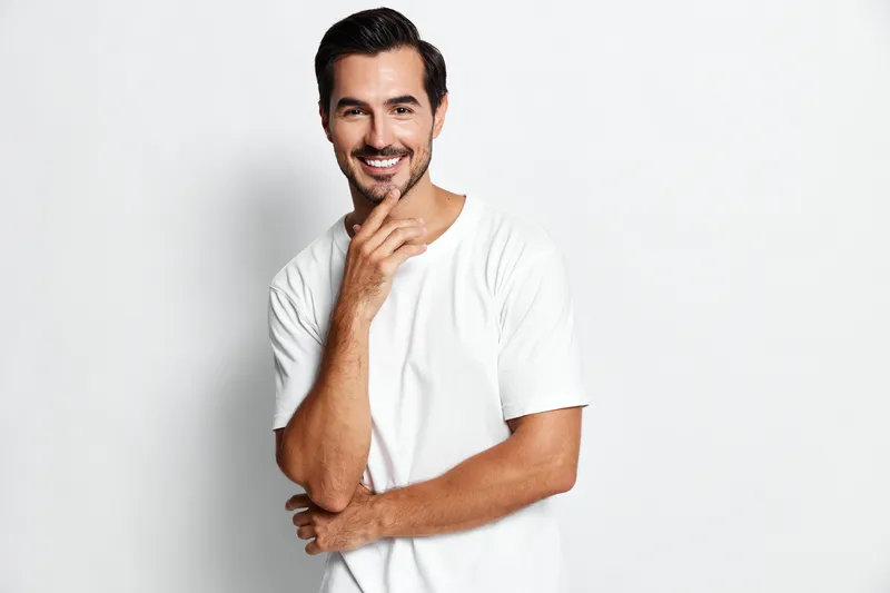 Smiling man in casual white t-shirt stands against a plain white background, looking confident and approachable, hand on chin, showcasing a relaxed vibe.