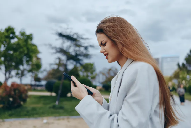 Confident businesswoman checking her phone outdoors in a professional outfit, expressing determination amidst a modern urban landscape with greenery.