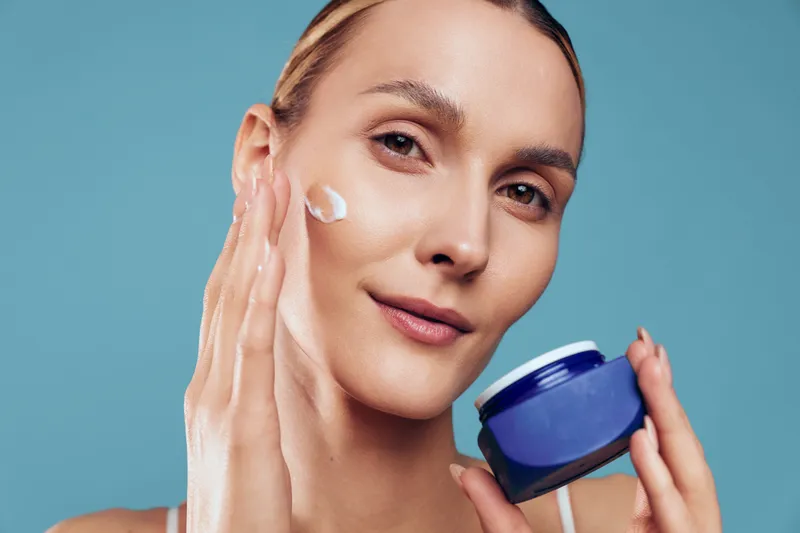Woman applying face cream on cheek holding cosmetic jar, skincare moisturizer routine for beauty, calm expression, close up against blue background, natural skin