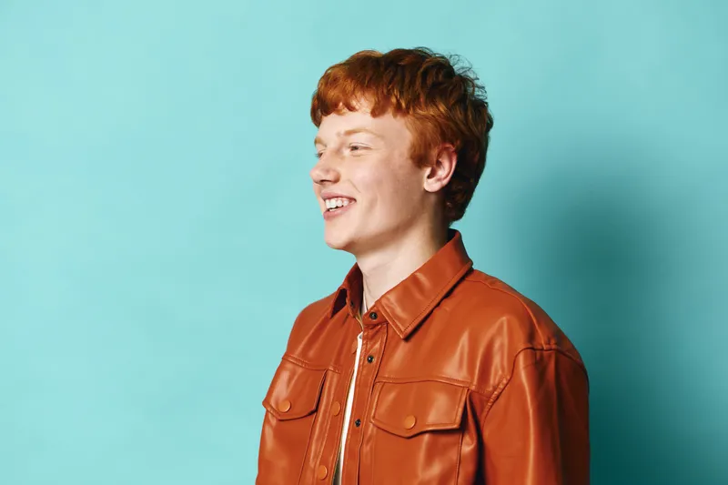 Young man with red hair wearing brown leather jacket smiling, looking aside on blue background in studio. Portrait of confident happy person in casual fashion clothing with natural lighting.