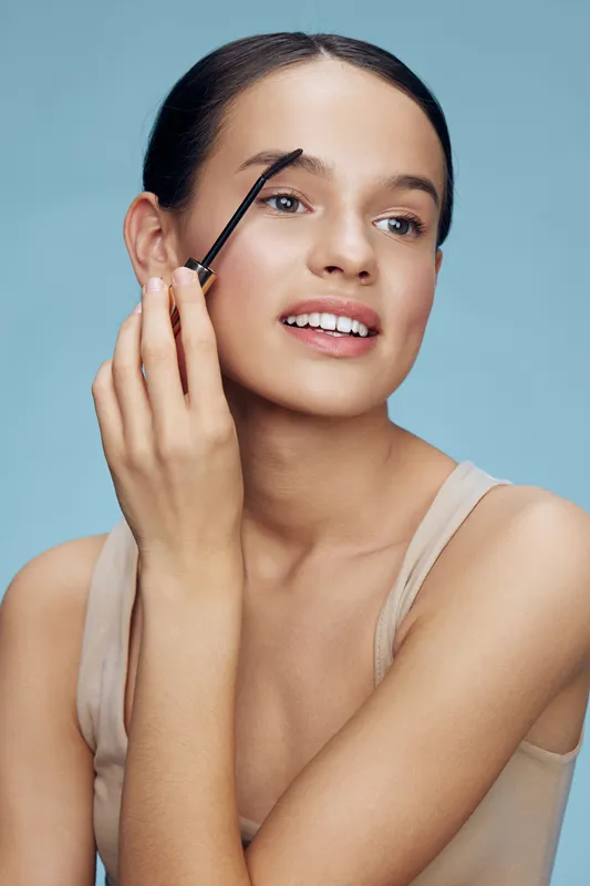 Young model applying eyebrow gel in a beauty routine with a soft smile, showcasing natural makeup against a pastel blue background, perfect for beauty advertising