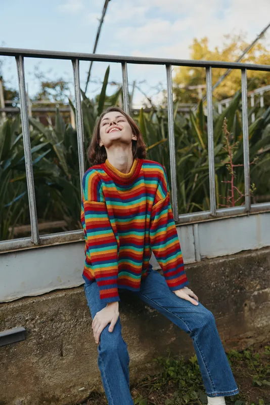 Young woman striped sweater sitting smiling jeans leaning back outdoor greenery, colorful knit and casual outfit on concrete ledge by metal railing for lifestyle fashion and travel.