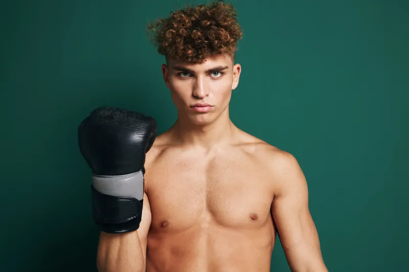 Young man with boxing gloves standing shirtless with focused expression isolated on green background. Boxer looks strong ready for training fight competition or workout session.