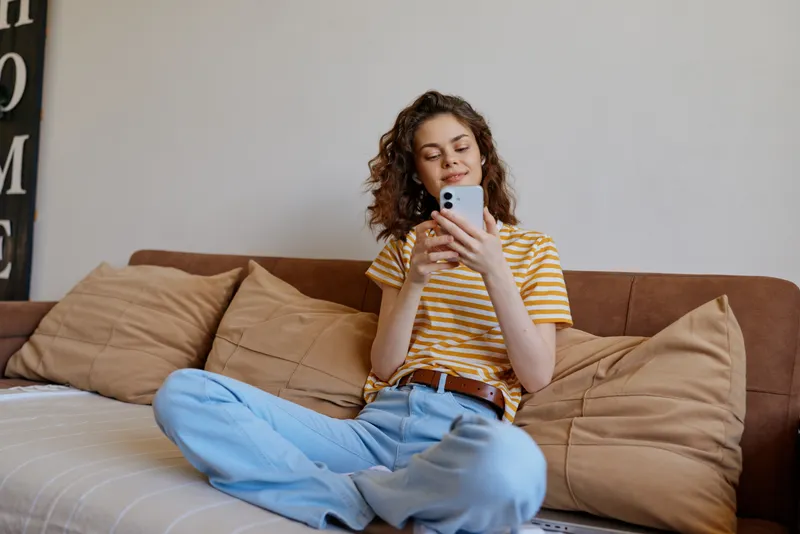 Casual woman sits on a sofa at home, scrolling on a smartphone, relaxed mood, natural light, striped shirt, blue jeans, cozy living room, candid lifestyle moment