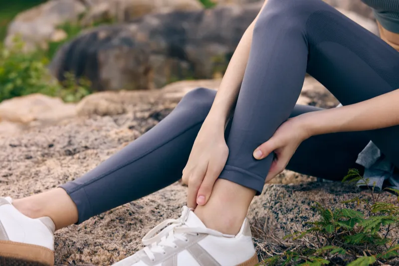 Young female in athletic wear experiencing ankle pain while sitting on a rock, showcasing the importance of injury prevention in fitness and outdoor activities