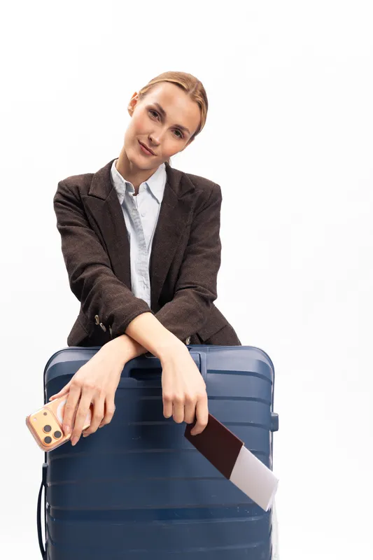 Young woman in brown blazer holds smartphone and passport, leaning on blue suitcase. White background highlights travel, business trip, and vacation concept with confident relaxed mood.