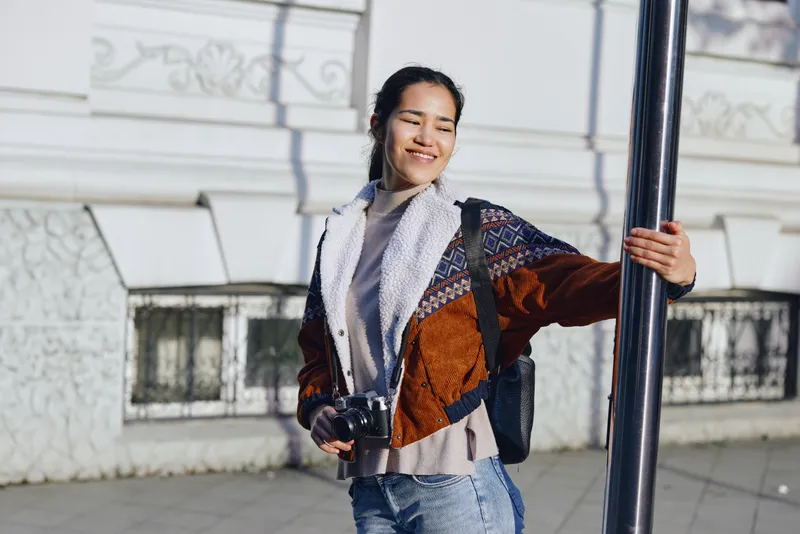 Smiling woman outdoors with camera, casual jacket and scarf beside a street pole, urban lifestyle moment, candid photography for travel, everyday joy and exploration