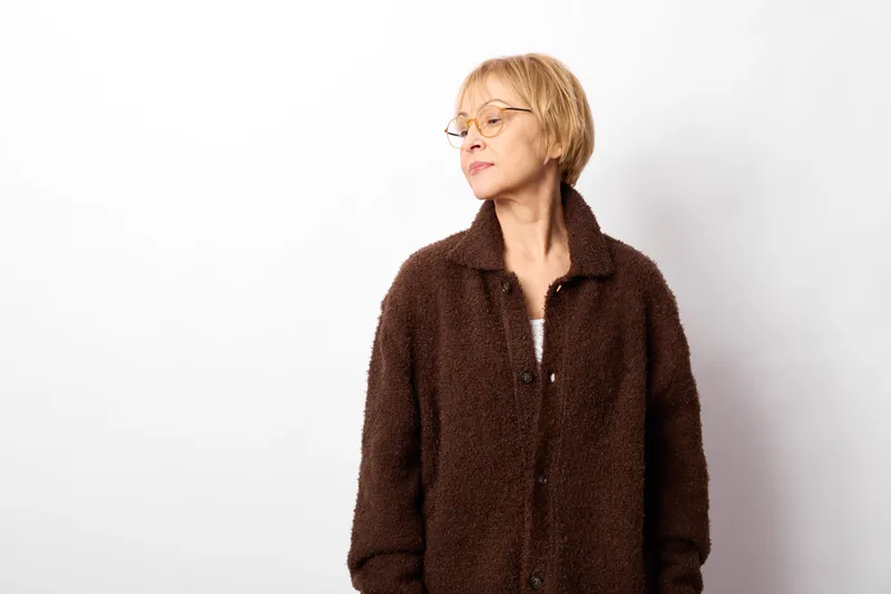 Senior woman with blonde short hair and glasses in a dark wool coat stands in a studio, looking away with thoughtful calm, concept of aging and maturity on white background