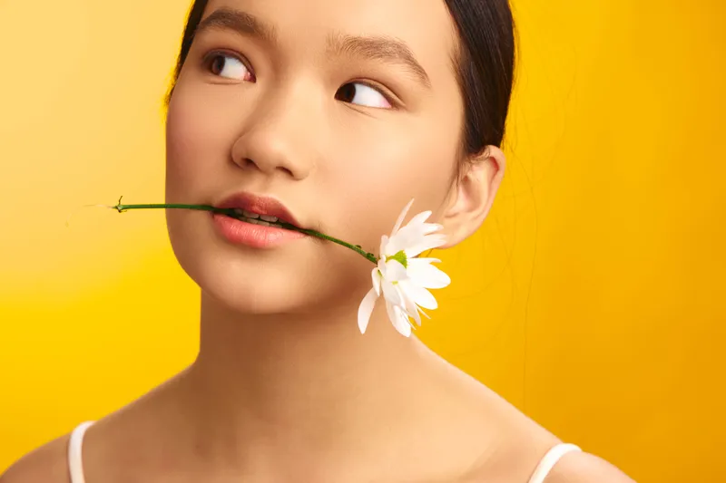 Asian woman gently holds a white flower between her lips, wearing a soft smile as she gazes off to the side. The bright yellow background creates a playful, artistic mood.