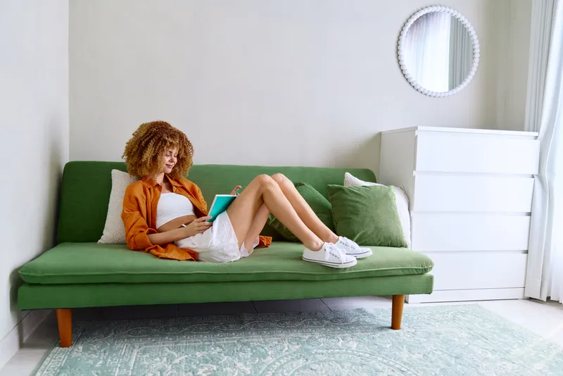Молодая женщина с вьющимися волосами отдыхает на зеленом диване, глубоко погрузившись в чтение книги