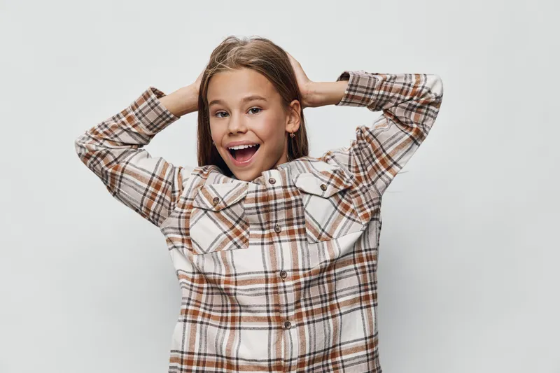 Girl with plaid shirt stands with hands behind head smiling and excited on light background. Happy child, casual outfit and joy with natural expression and youth energy.