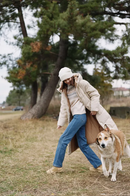 Woman walking a dog in a park wearing a beige coat jeans and beanie with calm autumn light tall trees and grassy field creating a relaxed outdoor lifestyle scene