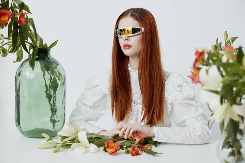 Stylish young model with long hair and futuristic sunglasses poses elegantly among fresh flowers in a bright and modern setting, showcasing contemporary fashion trends