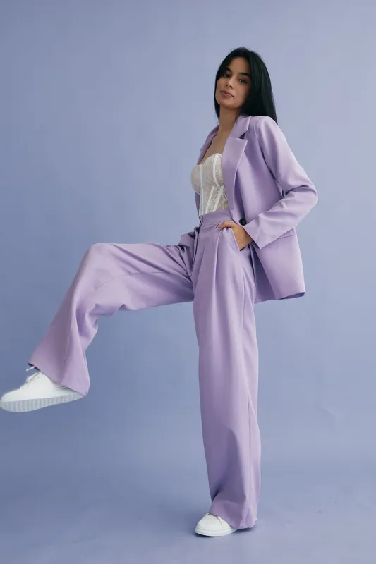 Stylish woman in lilac suit and white top posing confidently against a lavender background, showcasing modern fashion and minimalistic design in a studio setting.