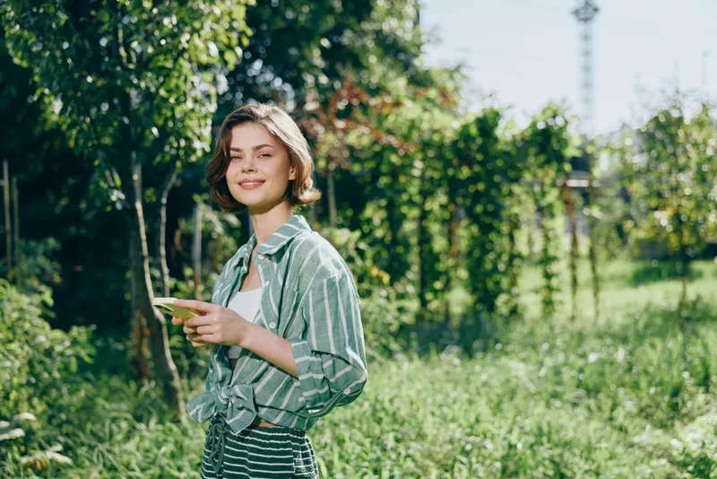 Young woman stands outdoors in a leafy garden, smiling as she holds a phone against a sunlit backdrop.