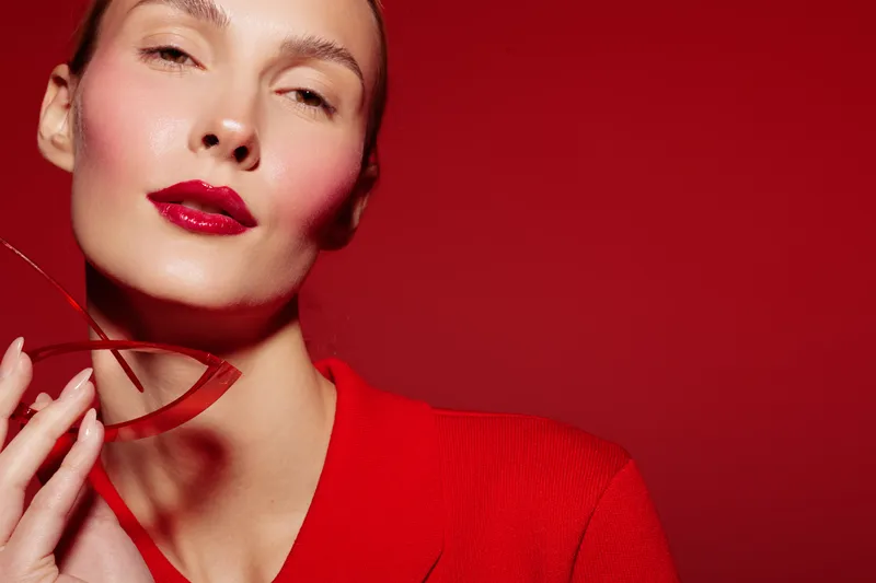 Close-up of a woman with red lipstick and red clothing holding a red ribbon against a red background. Concept of beauty, fashion, elegance, and monochrome style.