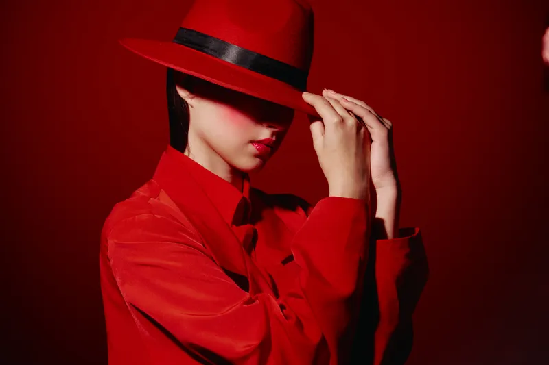 Fashionable woman wearing red silk shirt and matching wide brim hat poses against red background. Concept of beauty, style, creative fashion, elegance.