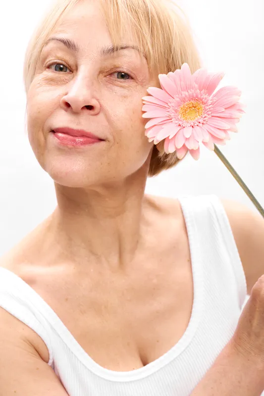 Senior woman smiling in closeup, holding a pink gerbera daisy flower on a stick, soft natural light, white background, gentle beauty portrait, calm mood wellness care