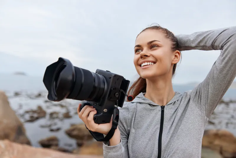 Smiling woman holding a camera by the seaside, wearing a gray hoodie, reflecting a joyful passion for photography Bright day, cool colors, outdoor adventure