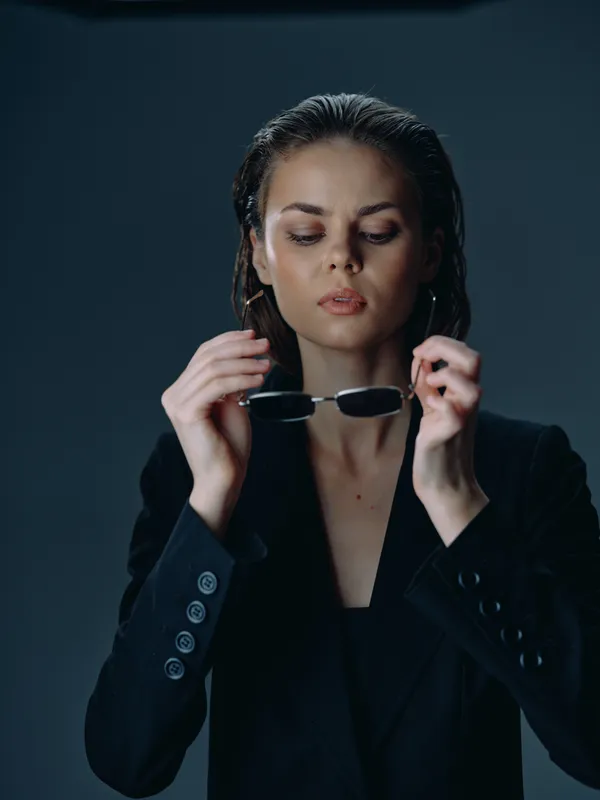 Young woman in black blazer holding sunglasses with wet hair, serious expression, studio portrait on dark background