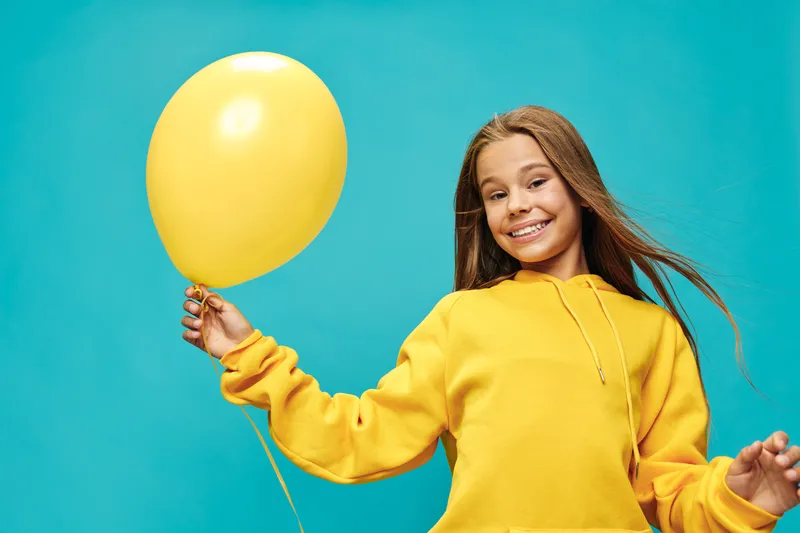 Girl smiling holding yellow balloon wearing matching hoodie against blue background, cheerful child portrait celebrating with bright colors and casual outfit indoors.