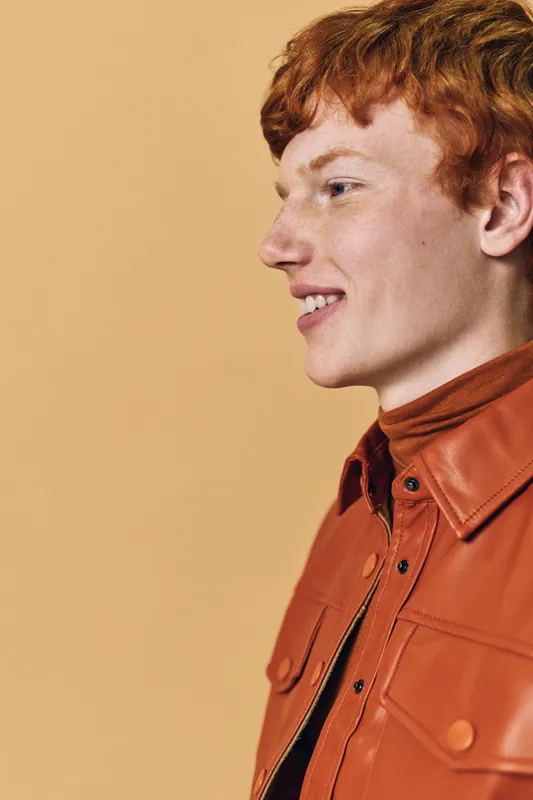 Young man with red hair smiling, wearing orange leather jacket and brown turtleneck, posing against plain beige background in studio portrait, side profile view.