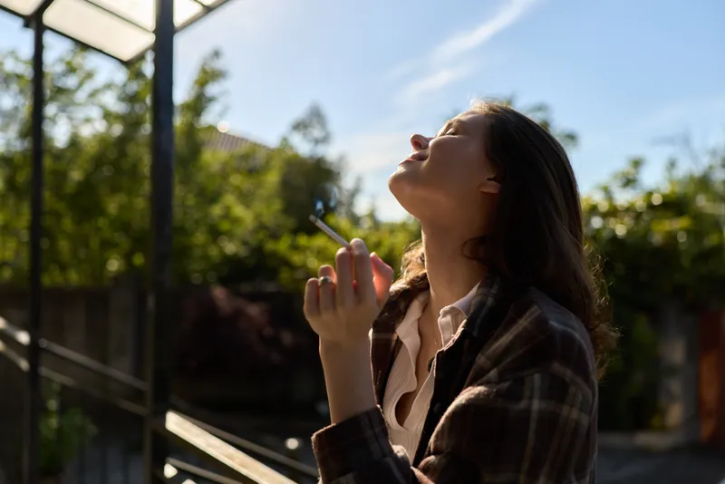 A woman stands outdoors on a sunny day, eyes closed, head tilted back, smoke rising near lips. Casual lifestyle moment capturing reflection, calm, and self care