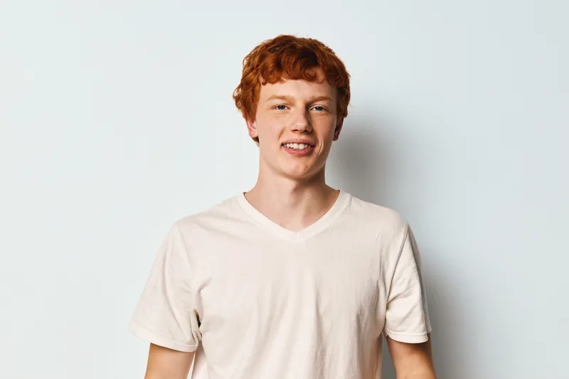 Young man with red hair wearing white t shirt standing and smiling on light background. Portrait of casual male model with freckled face and relaxed posture in studio setting.