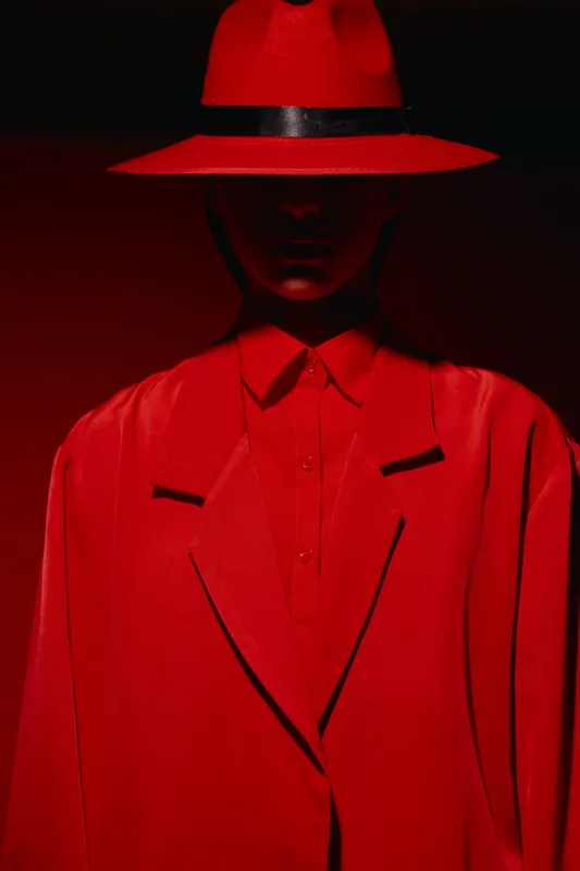 Fashion model in bold red hat and oversized coat in dark lighting. Creative beauty photo highlighting dramatic style and intense color contrast, studio portrait concept.