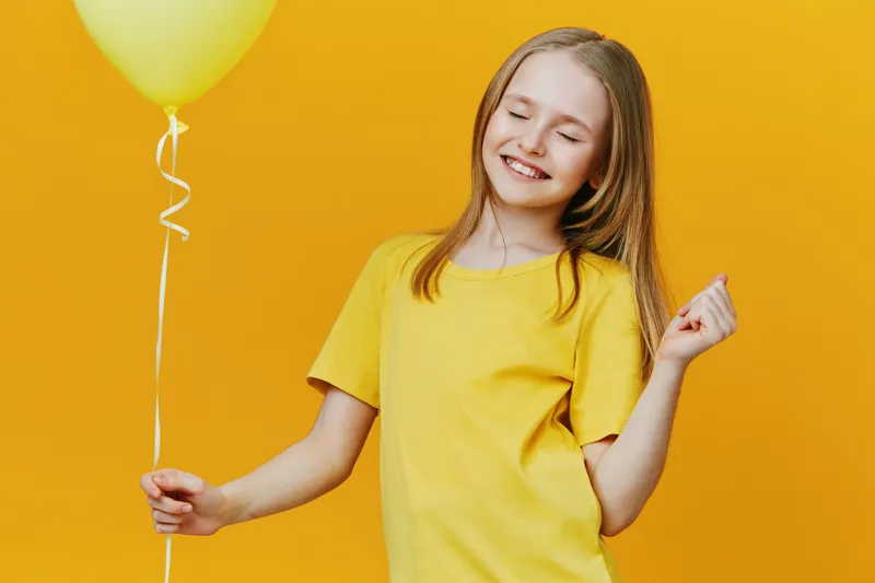 Happy girl with long hair in a yellow t shirt holding a yellow balloon against a vibrant yellow background, radiating joy and positivity