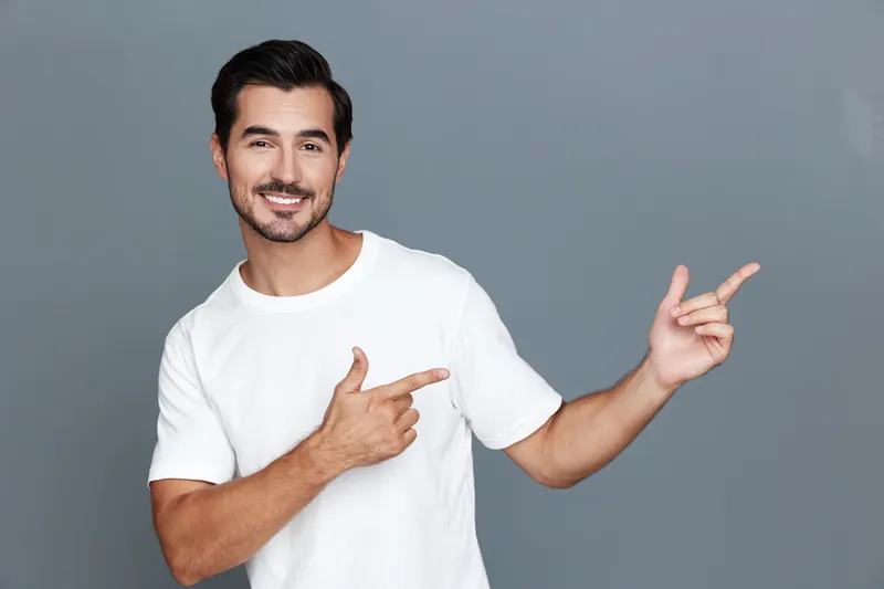Handsome smiling young man wearing a white t-shirt gestures with his hands while looking at the camera, showcasing a friendly and inviting personality against a solid gray background.