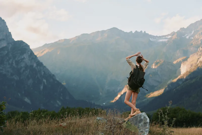 Woman hiking on a rocky outcrop in a mountain landscape, embracing outdoor adventure, carrying a backpack, under a bright sky, surrounded by vast nature and a sense of freedom