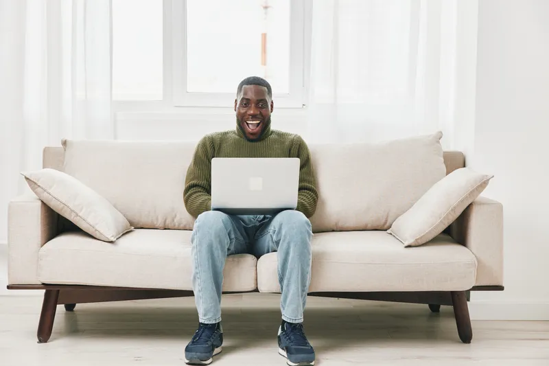 Happy Black man sitting on a couch with a laptop, wearing a green sweater and jeans, expressing joy and excitement in a bright, cozy living room