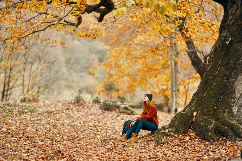 Женщина осенью в парке возле большого дерева, рядом с ней на земле лежит рюкзак