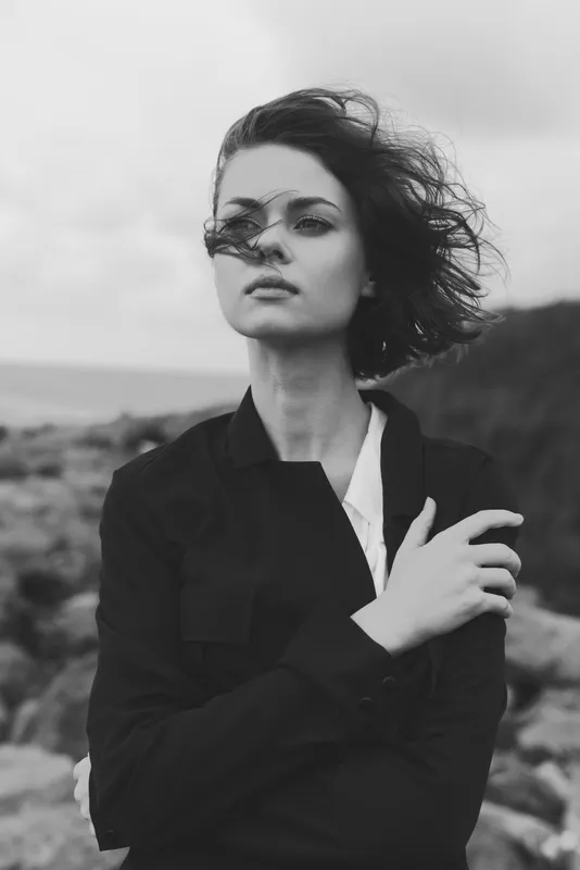 Monochrome image of a windblown woman in a dark jacket by a rugged shoreline, moody atmosphere, dramatic contrast and effortless style