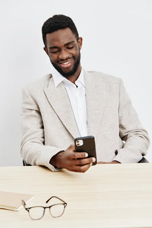 Smiling man using mobile phone, wearing beige blazer and white shirt, sitting at desk with eyeglasses and notebook, studio portrait, casual business lifestyle concept