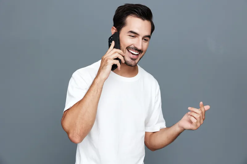 Smiling man in casual white t-shirt talking on mobile phone while looking happy at camera, isolated on grey background. Lifestyle and connection concept.