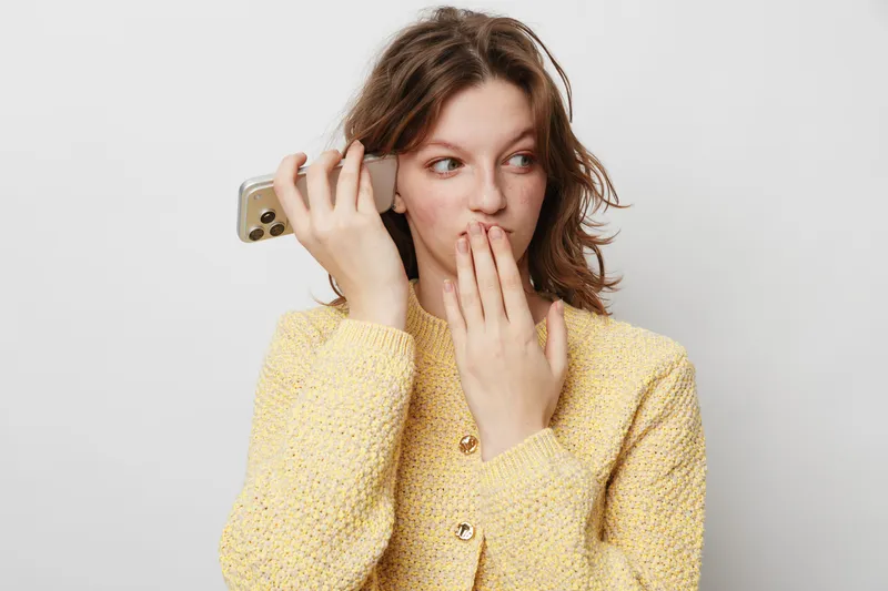 Young woman in yellow sweater holds smartphone near ear and covers mouth with hand, showing surprise or secret. Isolated on white background. Concept of communication, emotion, and privacy.