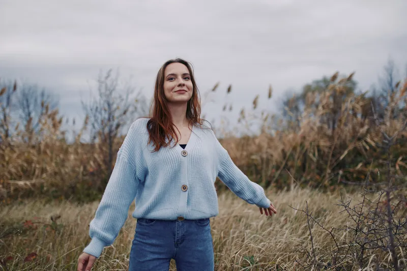 A young woman stands in a windswept meadow, smiling naturally with open arms, conveying quiet authenticity and credibility in a candid portrait highlighting genuine emotion and simple living.