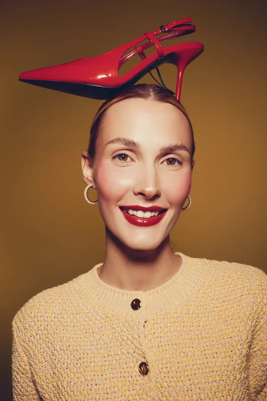 Smiling woman with red high heel shoe balanced on her head wears yellow knitted sweater and gold hoop earrings. Studio portrait with warm lighting and playful fashion concept.