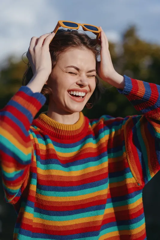 Woman smiling with sunglasses on head, colorful striped sweater portrait outdoors. Closeup of happy young female laughing with eyes closed, casual fashion and joy.