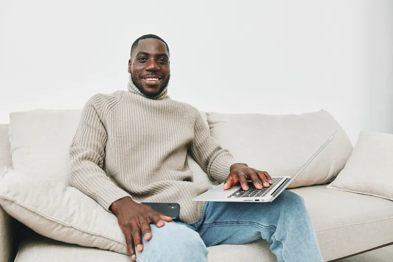 Smiling Black man working on a laptop at home in comfortable sweater during winter, showcasing home office vibes and cozy atmosphere