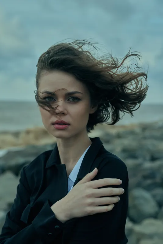 Windy coastal scene featuring a woman in a dark blazer, hair blown by wind, a moody yet calm mood, rocky shoreline and sea in the background, fashion vibe
