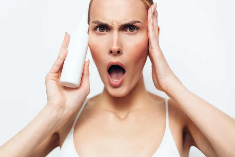 Surprised young woman holds white cosmetic bottle near face looks shocked touches head with hand isolated on white background skincare product advertisement concept.