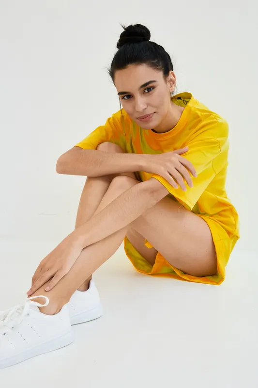 Young woman sitting on the floor in bright yellow dress and white sneakers, with hair in bun, relaxed smile, minimalistic white background and casual pose.