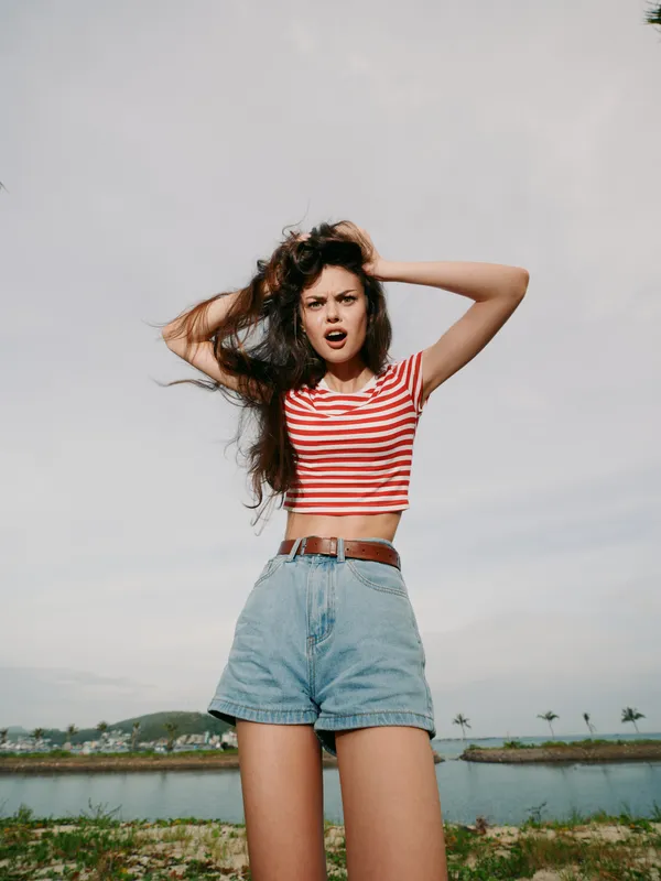 Energetic young woman stands outdoors in a striped crop top and denim shorts, striking a bold pose with wind in her hair against a bright coastal sky.