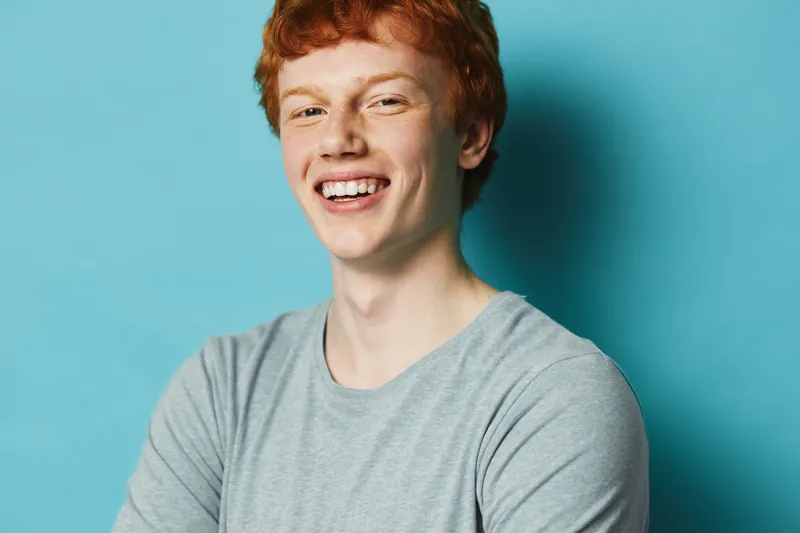 Young redhead man smiling with crossed arms wearing casual gray shirt standing against blue background for positive emotion and happiness portrait.