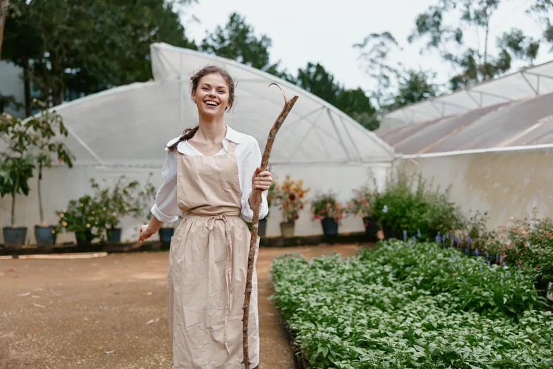 Happy young woman in an apron holding a stick in a greenhouse, surrounded by green plants, showcasing a joyful gardening spirit for seasonal nature lovers
