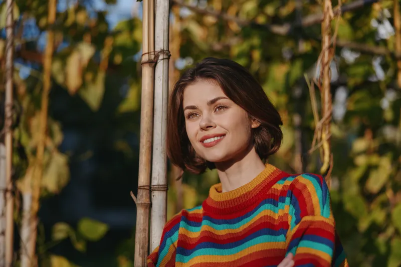 Woman smiling in colorful striped sweater outdoors, young adult leaning on a wooden pole among autumn foliage with short hair, casual outfit, warm sunlight lifestyle portrait for fashion and travel.